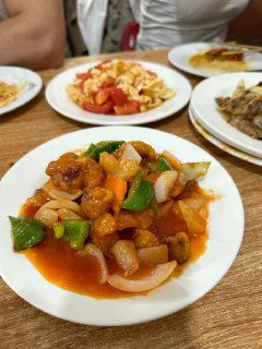 Порция китайского блюда в кисло-сладком соусе на деревянном столе, рядом видны тарелки с гарнирами — типичная подача в закусочной.