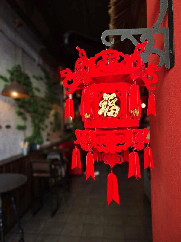 Крупный план красного китайского фонаря в интерьере кафе, на размытом фоне видны столы и растения — праздничный элемент оформления.