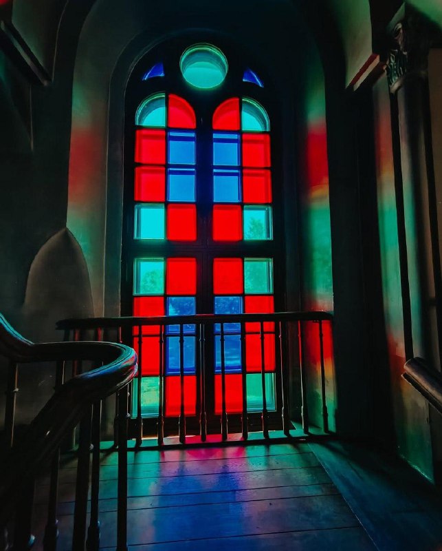 Арочное окно с цветными витражами и балконом позади — насыщенные красные и синие тона, свет, проникающий через стекло на пол.