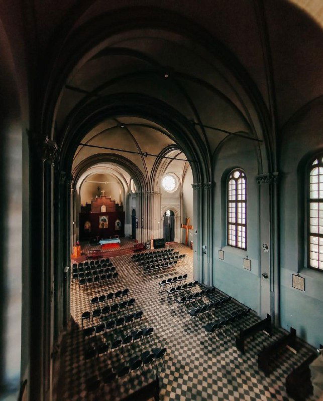 Панорамный вид на интерьер: высокий свод, ряды стульев, алтарь в глубине и характерная шахматная плитка на полу крупным планом.