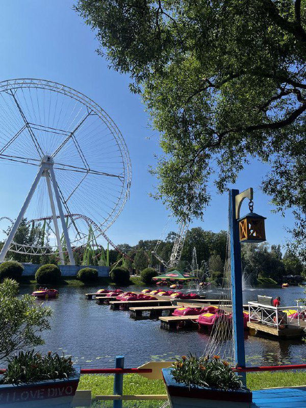 Озёрная зона парка с яркими прогулочными лодками на воде и крупным белым колесом обозрения на фоне чистого синего неба летним днём.