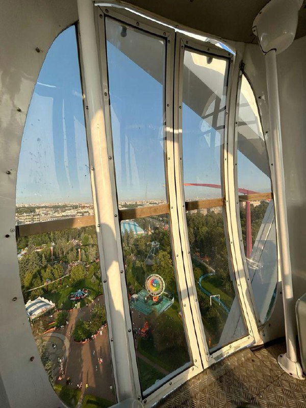 Вид изнутри кабинки колеса обозрения на парк развлечений и городскую панораму: стеклянные створки кабины и отражения летнего пейзажа.