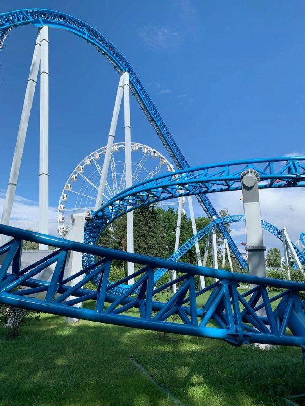 Крупный план синих рельсов американских горок на фоне белого кольца большого колеса обозрения и яркого голубого неба, вид внизу — трава.