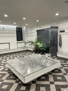 Белая кафельная комната с мраморной плитой, строгой геометрией пола и холодным светом — интерьер восстановленной банной зоны.