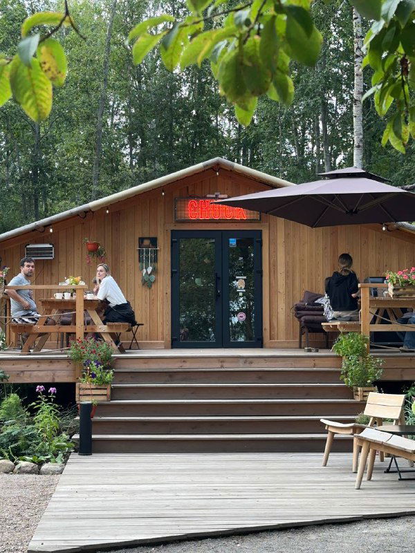 Деревянное здание ресторана Chuck на территории Lembo с летней террасой, столиками и зонтиками среди соснового леса и пешеходных дорожек.