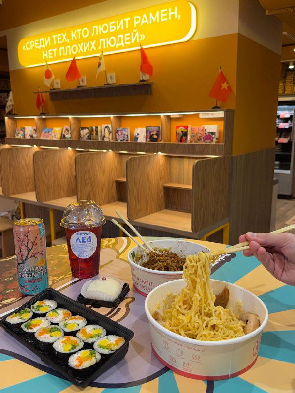 Интерьер: стол с двумя раменами, кимпабом и напитками на фоне уютной зоны с перегородками и неоновым текстом внутри KONO.