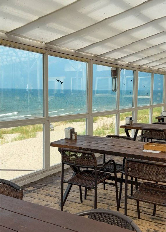 Интерьер прибрежного кафе с панорамными окнами и видом на море — столики рядом с огромными стеклянными панелями и пляжем за окном.
