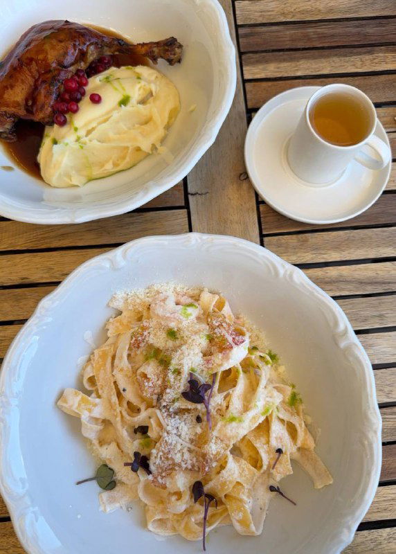 Два блюда: паста с пармезаном и мясное блюдо с пюре — сервировка ресторанного уровня на белых тарелках на деревянном столе.