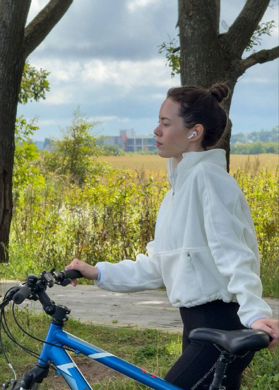 Девушка в спортивной кофте стоит рядом с велосипедом на фоне полей и деревьев — спокойный кадр прогулки по загородной дорожке у моря.