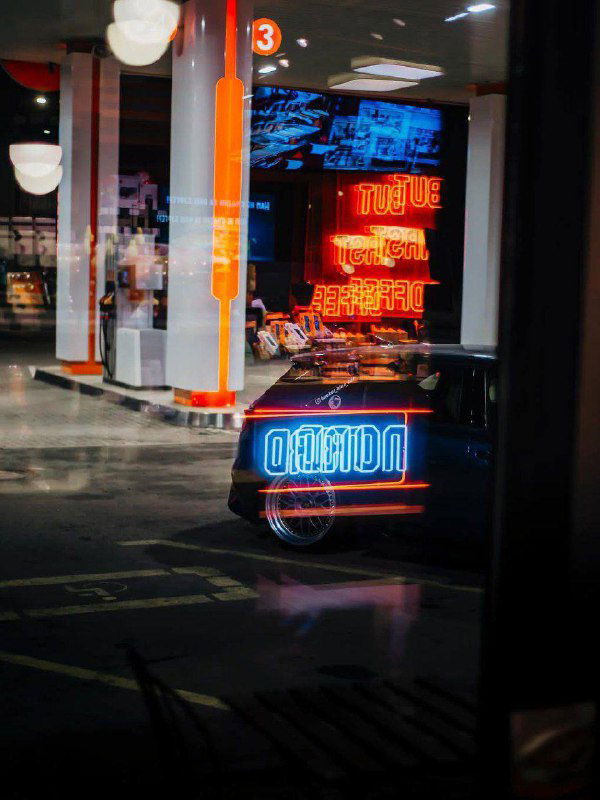 Отражение неоновой вывески и свет колонок на стекле — кадр с играми света и зеркальными отражениями в витрине заправки.