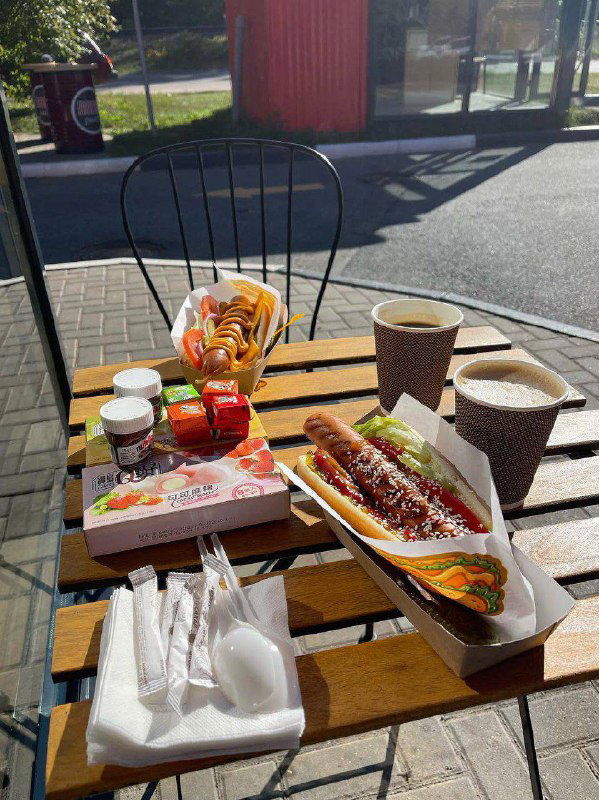 Дневной кадр: столик на улице у заправки с хот-догом, соусами и двумя стаканами кофе — пример небольшого пикника у точки обслуживания.