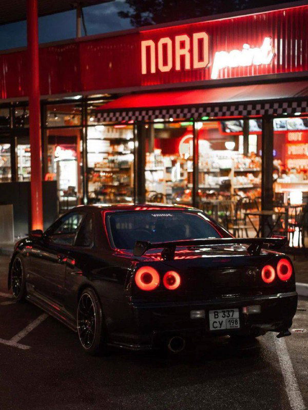 Ночной снимок с задней части спортивного автомобиля на фоне подсвеченного фасада Nord Point — контраст тёплого неона и аккуратной машины.