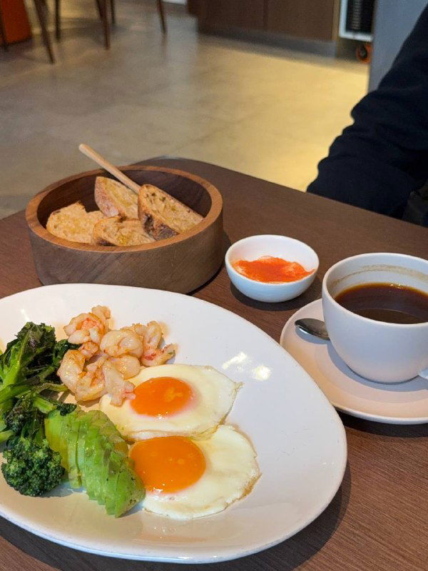 Завтрак с глазуньей, креветками, авокадо и брокколи рядом с соусом и чашкой кофе — сочетание для любителей сытных утренних блюд.