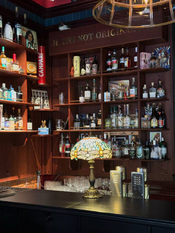Полки с алкоголем и витражная лампа на барной полке, интерьерная вывеска с названием и насыщенные тёмно-красные тона оформления паба.