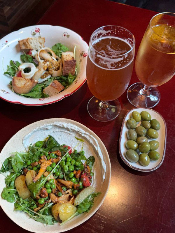 Два салата и бокалы с пивом и сидром: один салат с кальмаром и тёплыми гренками, другой с лисичками и горгонзолой; рядом небольшая тарелка оливок — стол с закусками.