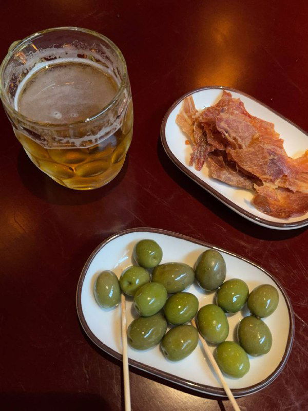 Подача к пиву: тарелка зелёных оливок и тарелка чипсов парманелло рядом с бокалом пива на деревянном столе — простая закуска для напитков.