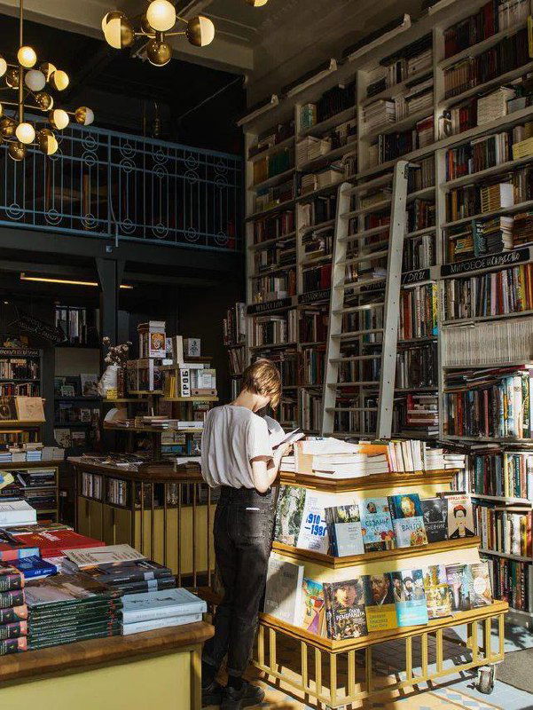 Высокие деревянные стеллажи, галерейный балкон и человек у прилавка — интерьер просторного книжного магазина с естественным освещением.