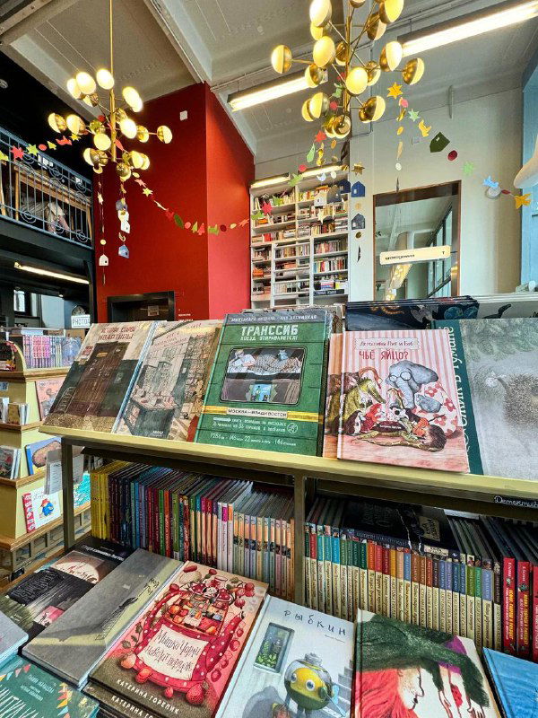 Прилавок с выставленными художественными и детскими изданиями на фоне полок; крупный план обложек и подборок в магазине.