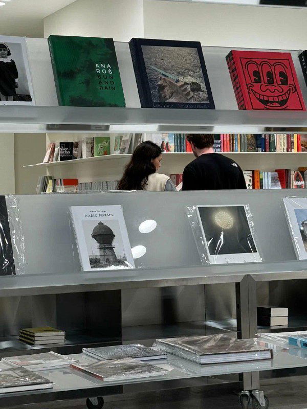 Полка с арт‑и дизайн‑книгами и двое посетителей, выбирающие издания; современный минималистичный стеллаж и светлый интерьер магазина.