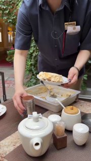 Официант подаёт чайную сервировку: белый чайник, чашки и блюдца — кадр из сервиса на летней веранде кафе.