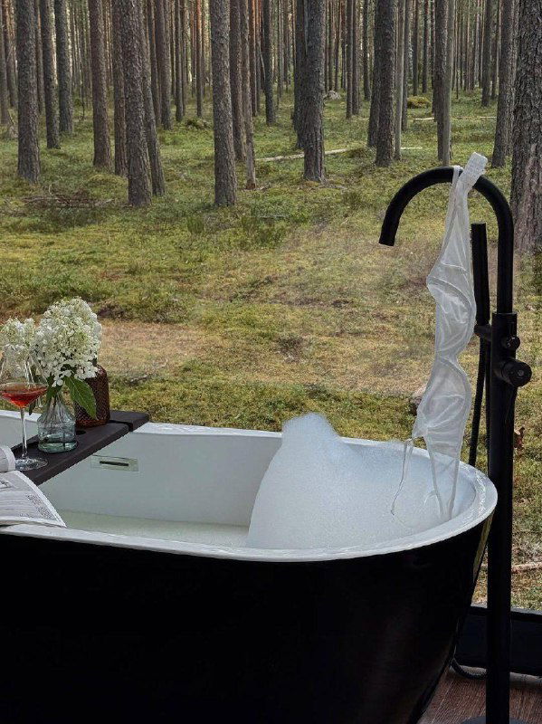 Ванна на открытом воздухе с обильной пеной стоит посреди хвойного леса — атмосферный кадр глэмпинга с видом на моховой покров и высокие сосны.
