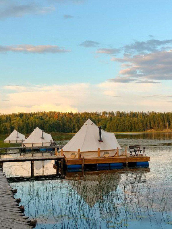 Плавающие шатры и домики на понтонах у берега озера на закате — тенты в форме типи, рефлексы воды и лес на горизонте создают романтичную картину.