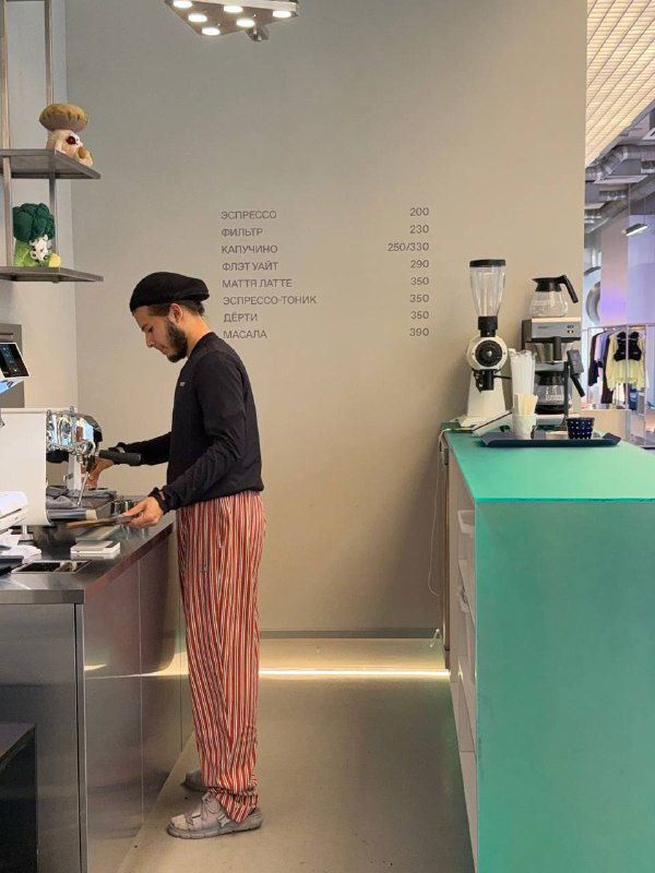 Бариста в кепке готовит кофе за профессиональной кофемашиной в IRNBY; на стене минималистичное меню и видна рабочая зона.