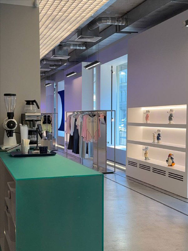 Интерьер магазина IRNBY: кофейная стойка на первом плане, за ней вешалки с одеждой и витрины с предметами дизайна в светлом пространстве.