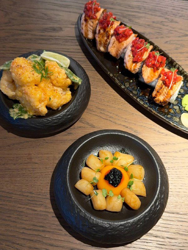 Три тёмные керамические тарелки с закусками: обжаренные гребешки, креветки в кляре и небольшие порции на деревянном столе в TAO