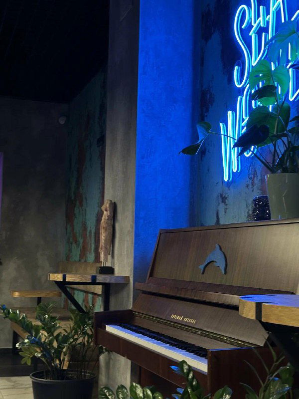 Угол интерьера SeaWolves: старинное пианино на фоне фактурной стены, растительность и неоновая подсветка создают морскую атмосферу заведения.