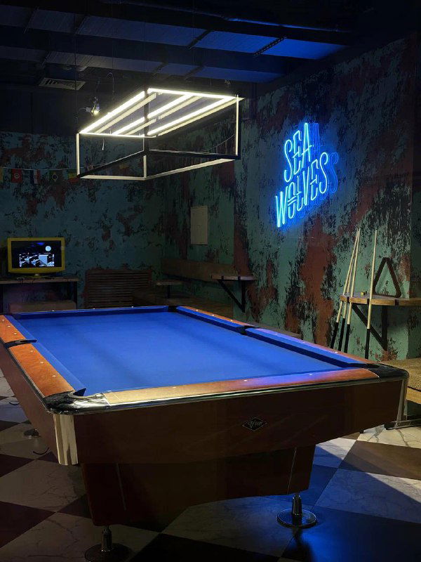 Бильярдный стол с синим сукном и логотипом SeaWolves на стене; кадр показывает игровой стол и часть просторного зала в баре-клубе.
