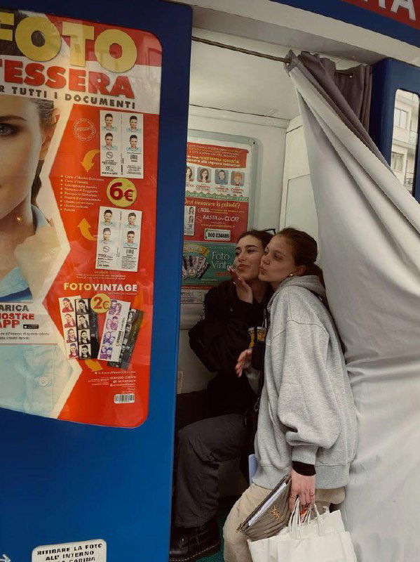 Две девушки позируют в уличной фотокабине с поднятой шторкой; рядом яркая рекламная панель с надписью FOTO — уличная сцена съёмки.