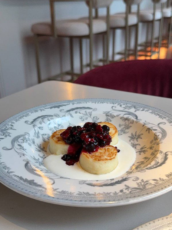 Сырники со сметаной и ягодным вареньем на белой тарелке, рядом чайная чашка — фокус на текстуре и соусе.