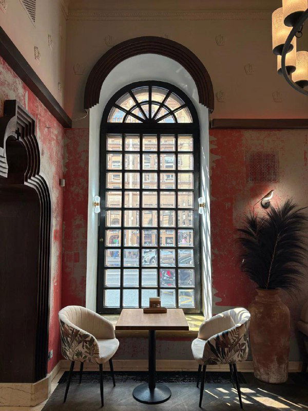 Небольший столик у большого арочного окна в кафе: свет, кресла и расслабленная атмосфера для пар и одного человека в Петербурге.