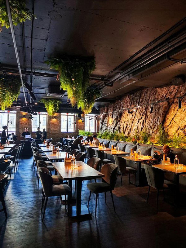 Просторный зал ресторана с высокими потолками, зелёными растениями и длинными рядами столов — формат для больших компаний и вечеров.