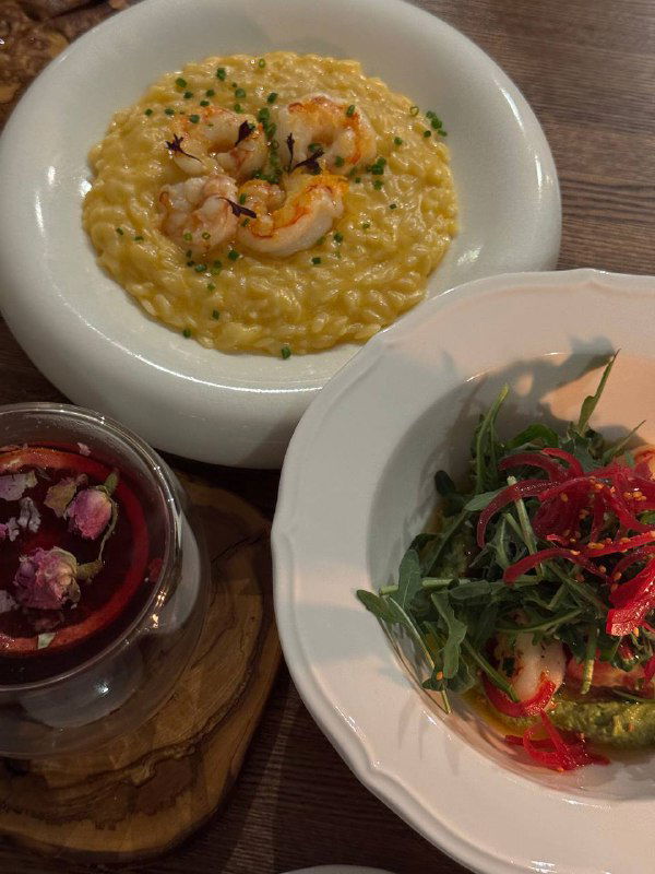 Тарелка с орзо и крупными лангустинами в кремовом соусе рядом с салатом из свежих томатов и бокалом пряного напитка.