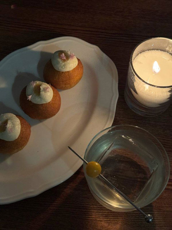 Небольшая тарелка с фирменными пончиками и бананом рядом со свечой и прозрачным бокалом с коктейлем на деревянном столе.