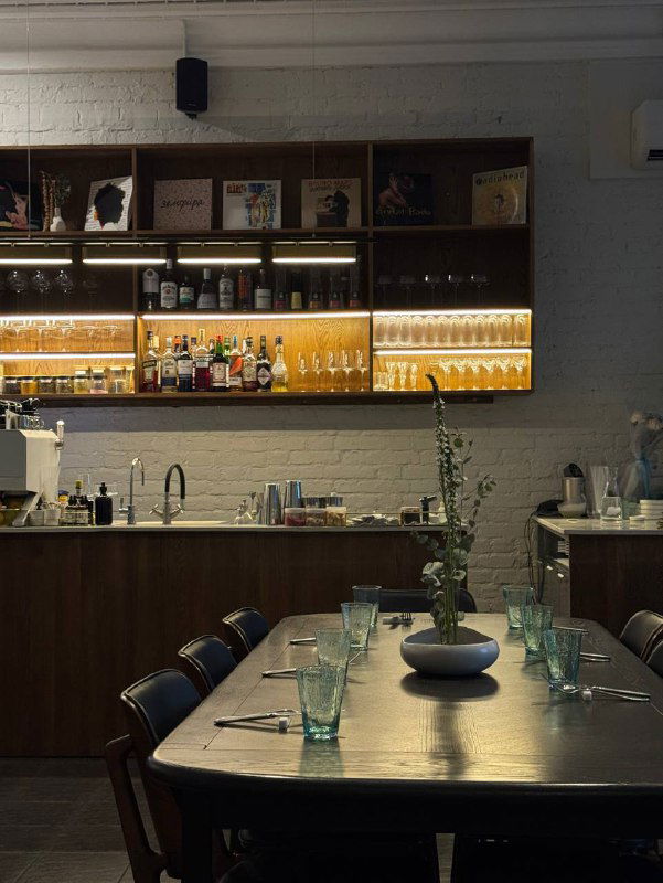 Длинный стол с сервировкой, полками с бутылками на фоне и минималистичным освещением — дневной или вечерний кадр бара Soul Babe.