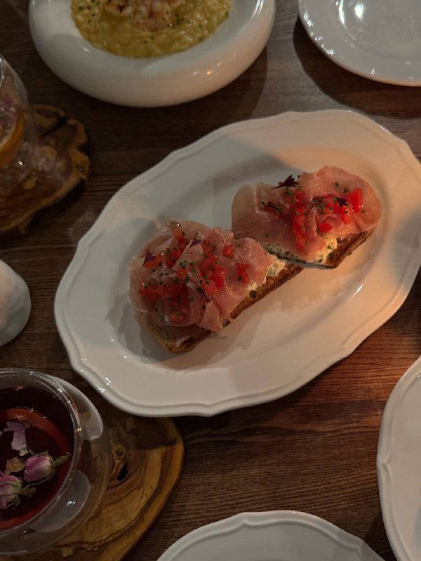 Брускетты с томатной сальсой и тонкими ломтиками ветчины на белой овальной тарелке, рядом видна часть орзо и салата.