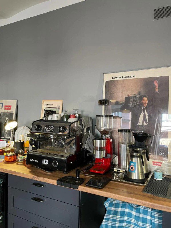 Барная зона с профессиональной кофемашиной, кофемолками и аксессуарами на фоне постера — рабочее место кофейного островка в магазине-кофейне.