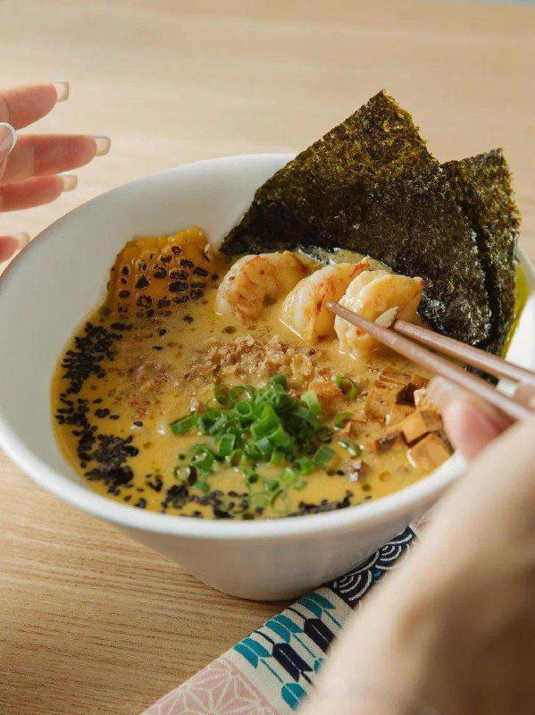 Крупный план горячего рамёна: миска с бульоном, нори, креветками, зелёным луком и палочками в руках — аппетитный вид традиционного блюда.