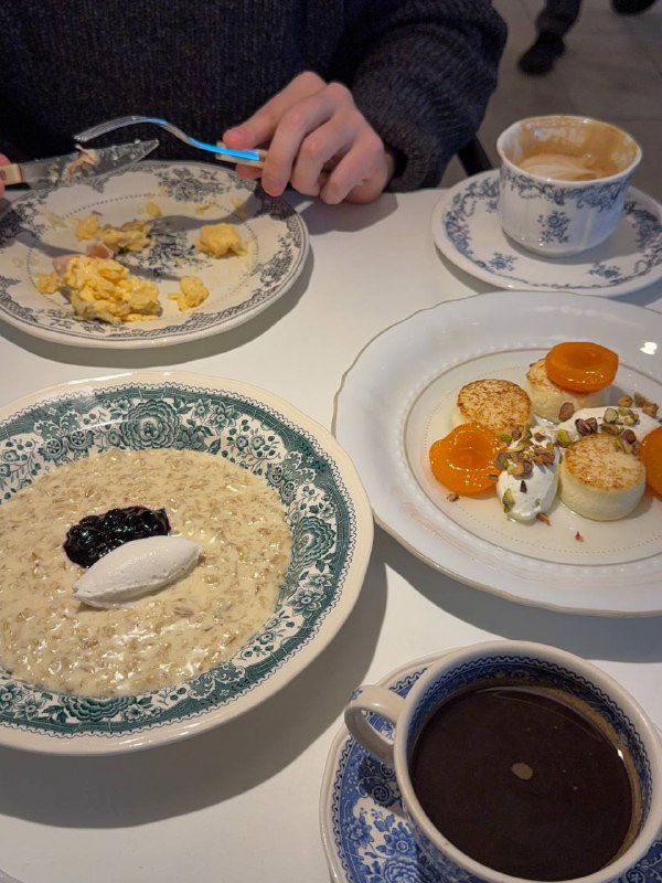 Несколько блюд на столе: овсянка на кокосовом молоке с конфитюром, сырники и напитки на фоне сервировки в бистро.