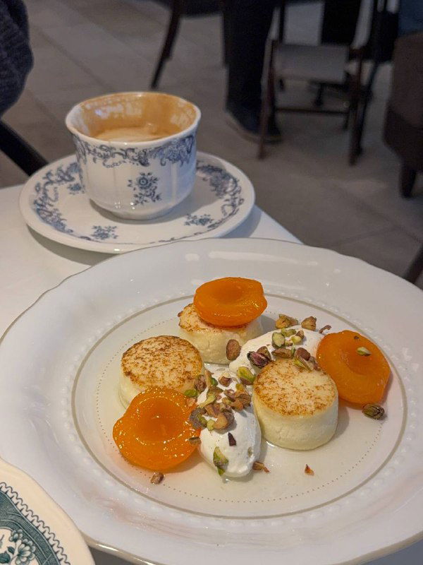 Крупный план сырников с рикоттой — подача с абрикосами, фисташками и ванильным кремом на белой тарелке, акцент на текстуре и украшениях.