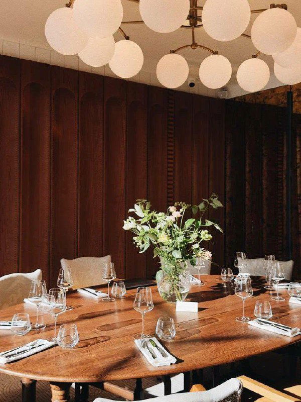 Сервированный стол в бар-ресторане Cinéma Michèle: деревянный стол, приборы, бокалы и ваза с цветами на фоне тёмной деревянной стены и люстр.