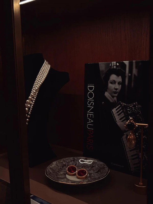 Витрина с украшениями и художественной книгой: тёмный ретро-декор, жемчужные аксессуары и тарелочка с деталями оформления внутри заведения.