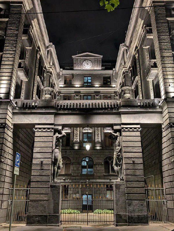 Ночное или раноутреннее освещение арки доходного дома Роберта Веге: подсветка фасада, видны колонны, атланты и центральный ризалит с деталями.