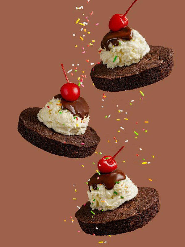 Несколько шоколадных десертов с кремом, вишней и посыпкой на коричневом фоне, художественная «парящая» компоновка для подачи.