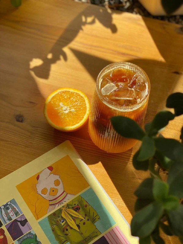 Солнечный натюрморт на столике в книжном: стакан с апельсиновым напитком, половинка апельсина и открытая книга — тёплая утренняя композиция для чтения