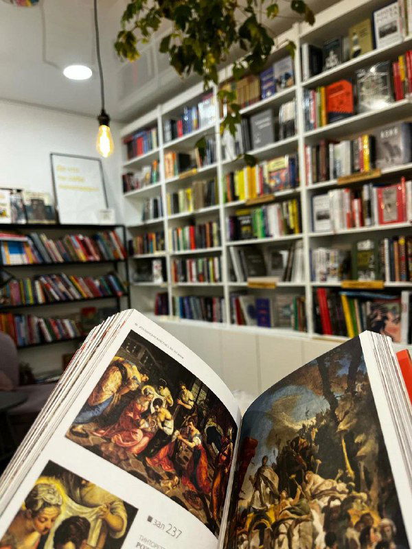 Крупный план открытой книги с репродукциями картин на фоне широких стеллажей — фокус на печатной культуре и ассортименте магазина