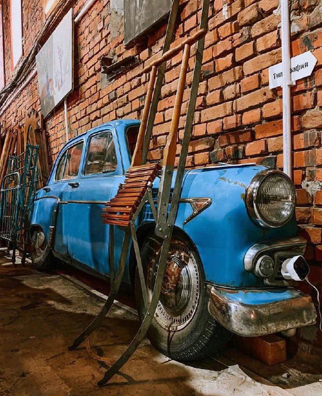 Синий ретро-автомобиль у кирпичной стены внутри магазина, окружённый аксессуарами и лестницей — атмосферный реквизит для фотосета.